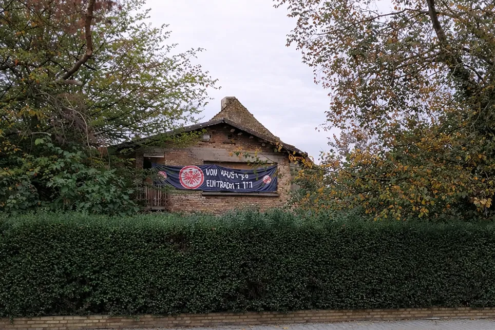 Ein altes Backsteingebäude hinter einer Hecke und zwischen Bäumen.