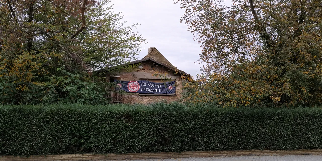 Ein altes Backsteingebäude hinter einer Hecke und zwischen Bäumen.